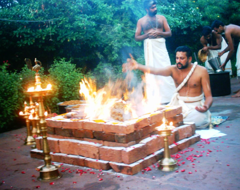 A Hindu priest performing a ritual fire ceremony, seated in front of a brick altar surrounded by flames, with other participants in traditional attire in the background.