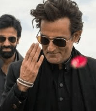 A man wearing sunglasses and a black outfit, greeting with a hand gesture, with another man smiling in the background.